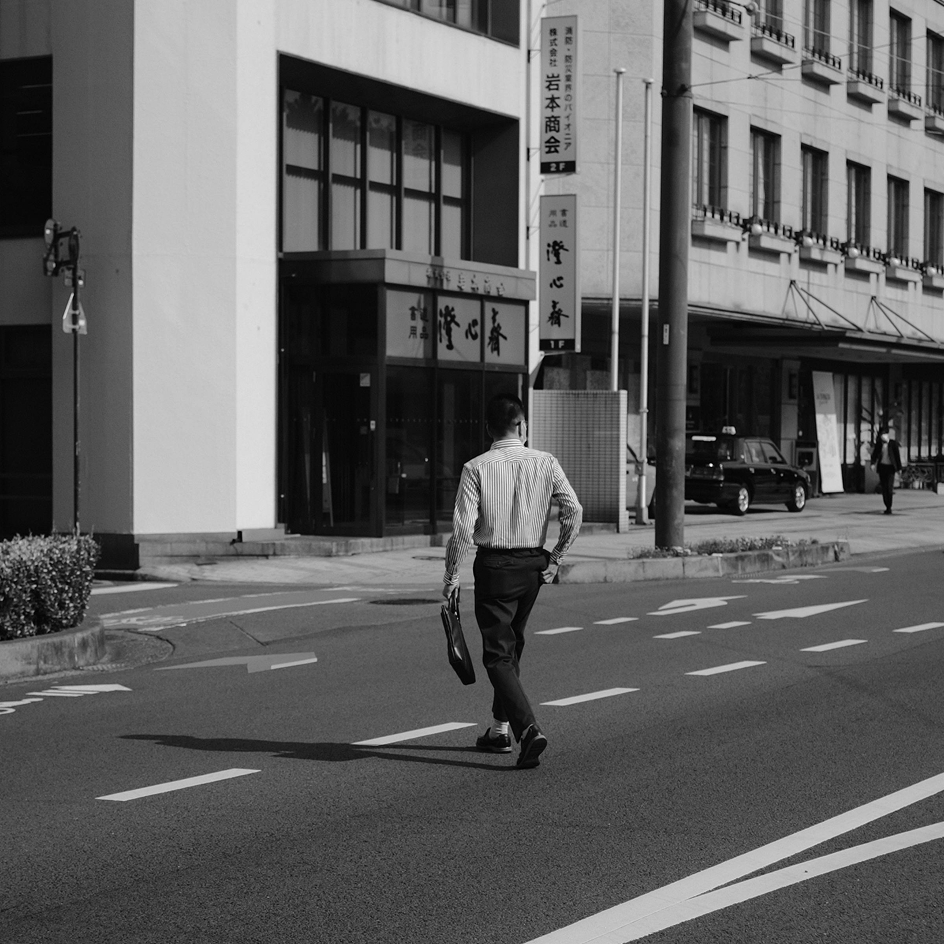 Man in striped shirt carrying a briefcase walking across a city street in front of buildings with Japanese signage.