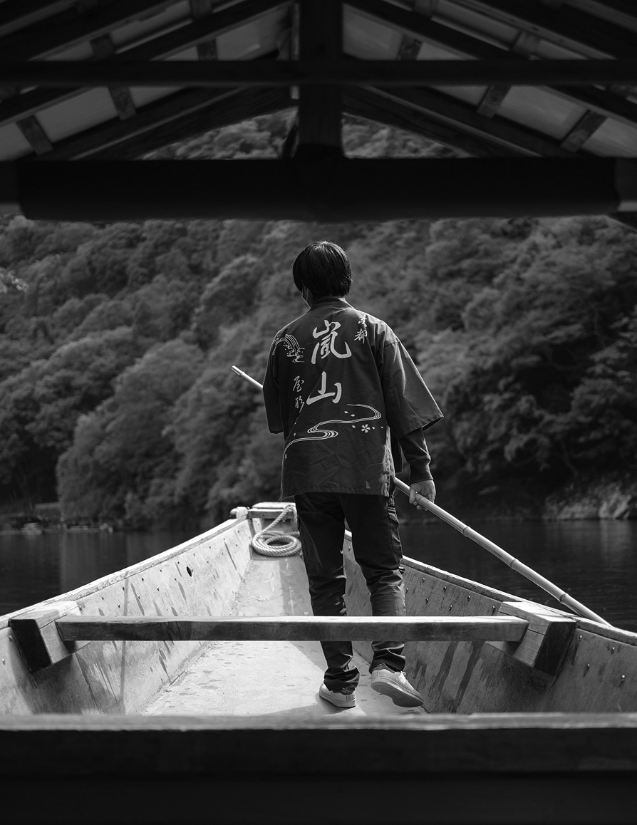 Person standing at the back of a wooden boat holding a long pole, navigating on a calm river with forested hills in the background.