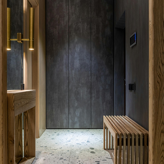 Modern hallway with terrazzo floor, textured dark gray walls, wooden bench, and vertical brass light fixtures next to a mirror.