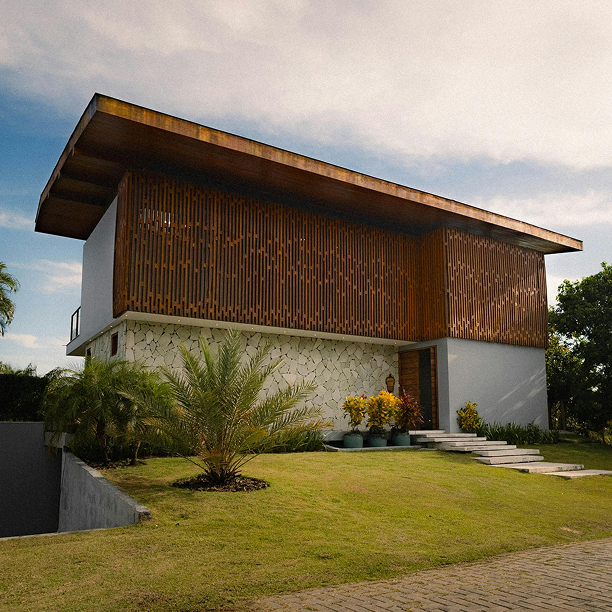 Modern two-story house with a large overhanging roof, wooden lattice on the upper level, stone textured lower wall, and landscaped yard with palm plants.
