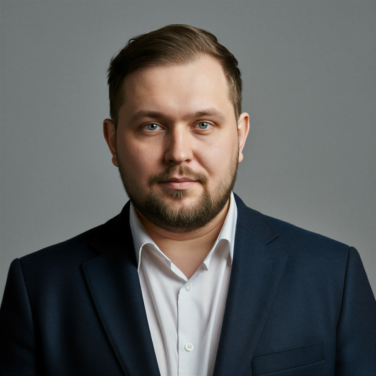 Portrait of a bearded man with blue eyes wearing a dark suit jacket and white shirt against a gray background.