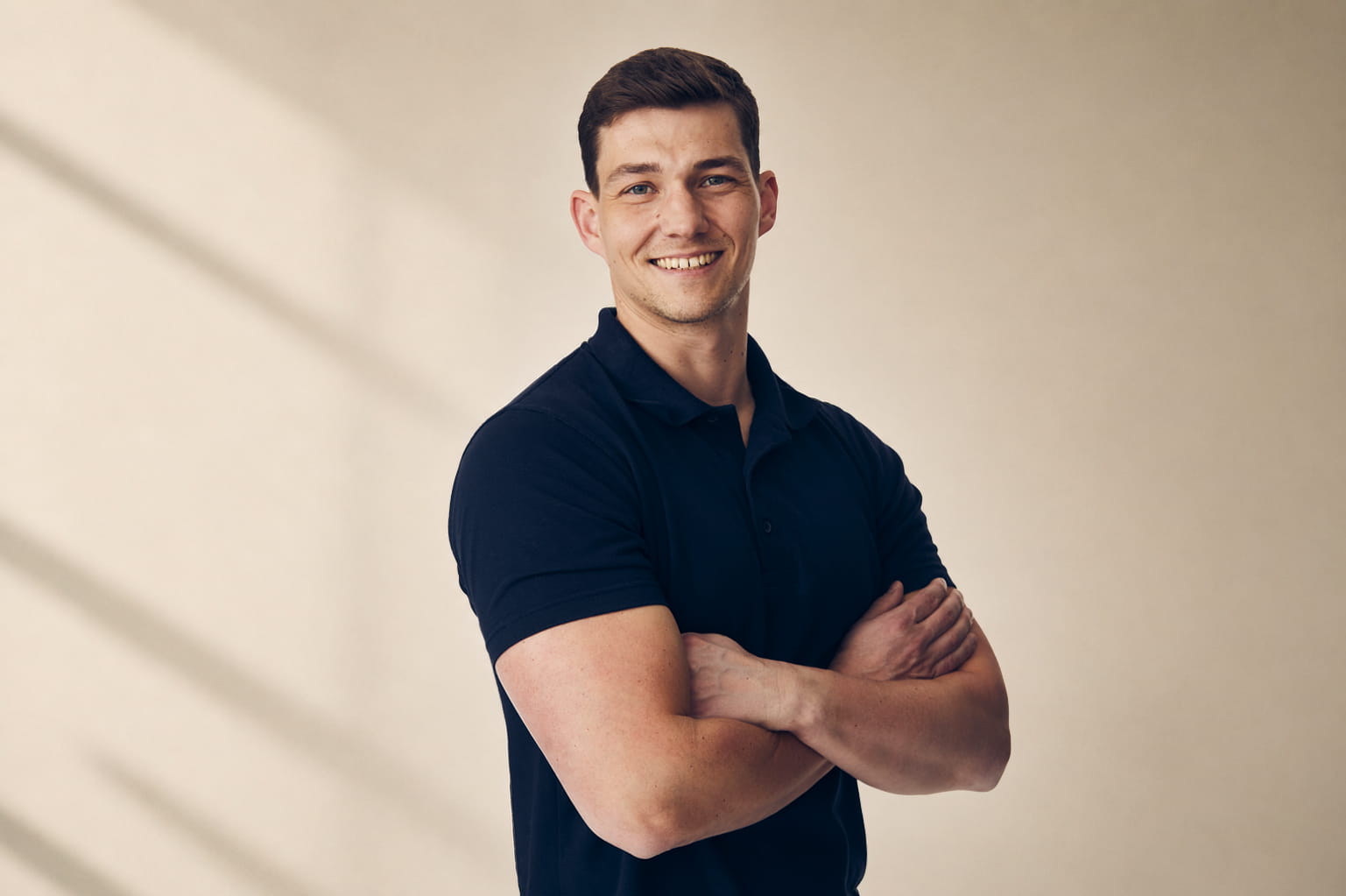 Smiling man with short dark hair wearing a black polo shirt, arms crossed against a light background.