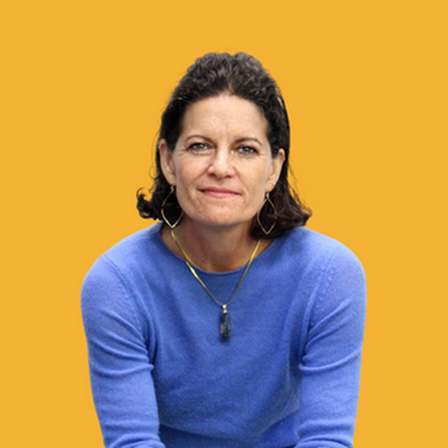 Woman with dark hair wearing a blue sweater and pendant necklace against an orange background.