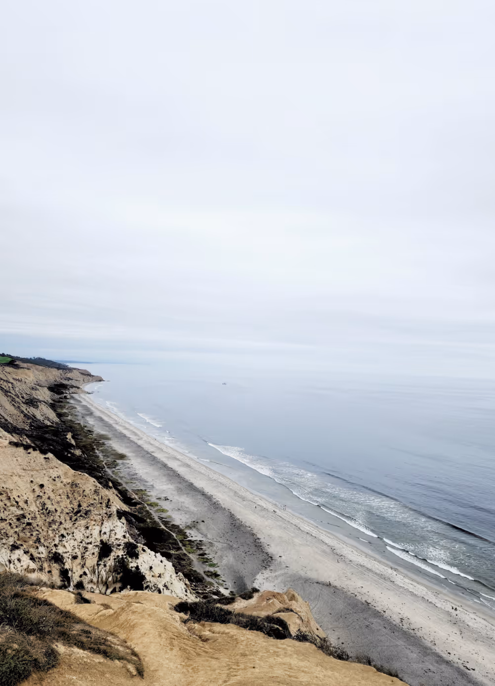 Aerial view of a long sandy beach with gentle waves and cliffs on a cloudy day.