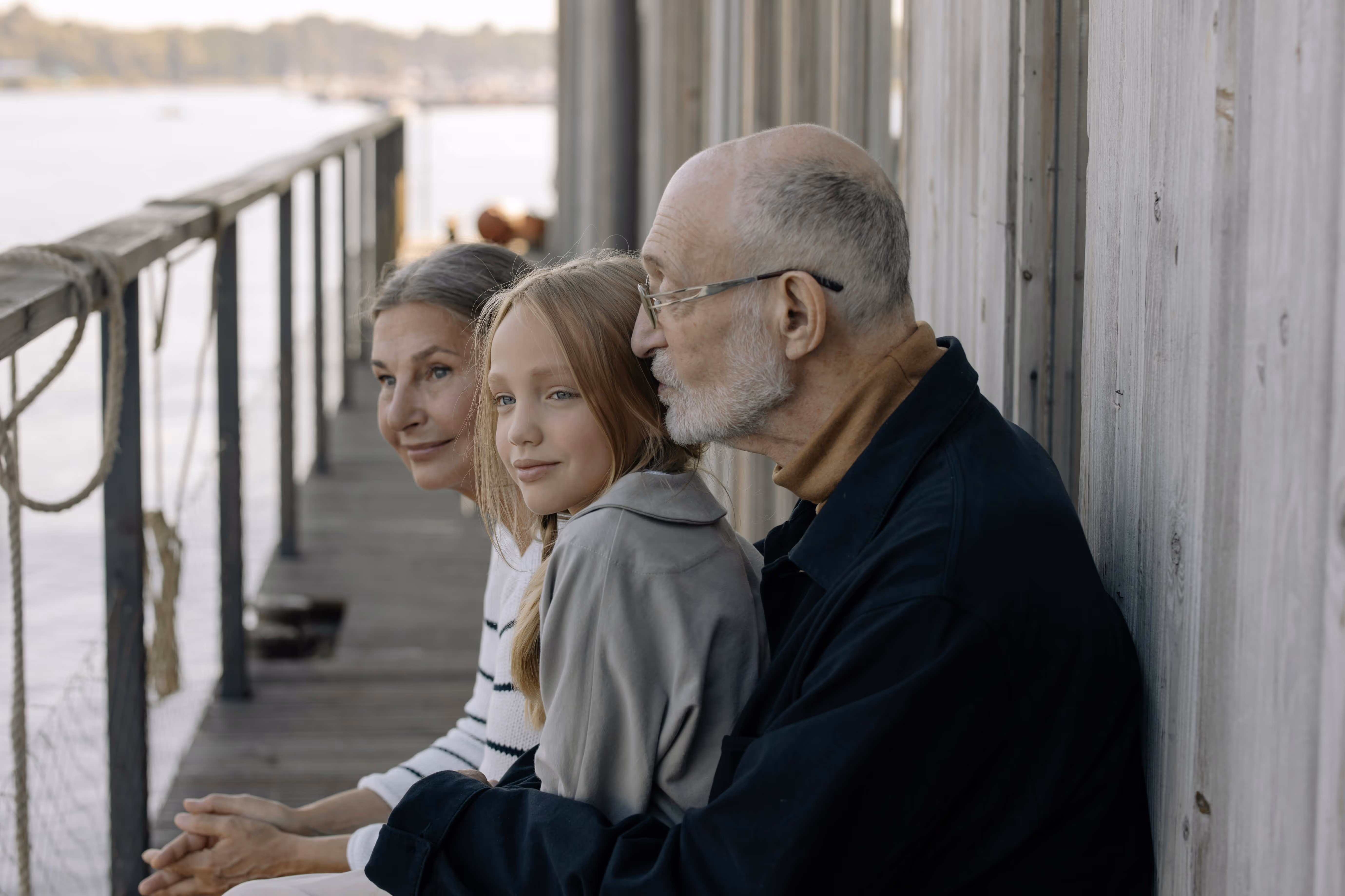 Three generations of a family sitting side by side on a wooden dock by the water, looking ahead peacefully.