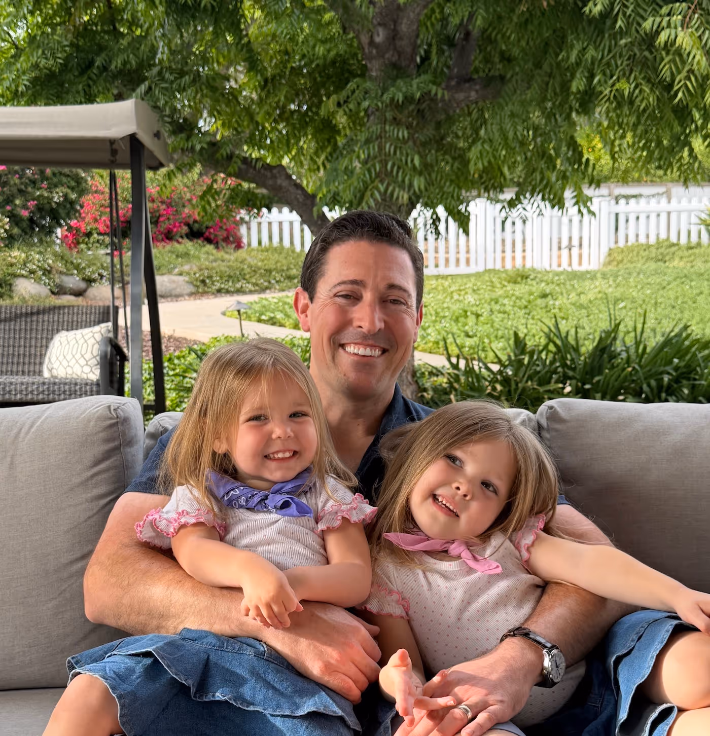 Smiling man sitting on an outdoor couch holding two young girls wearing white tops with pink trim and denim skirts, with greenery and a white picket fence in the background.