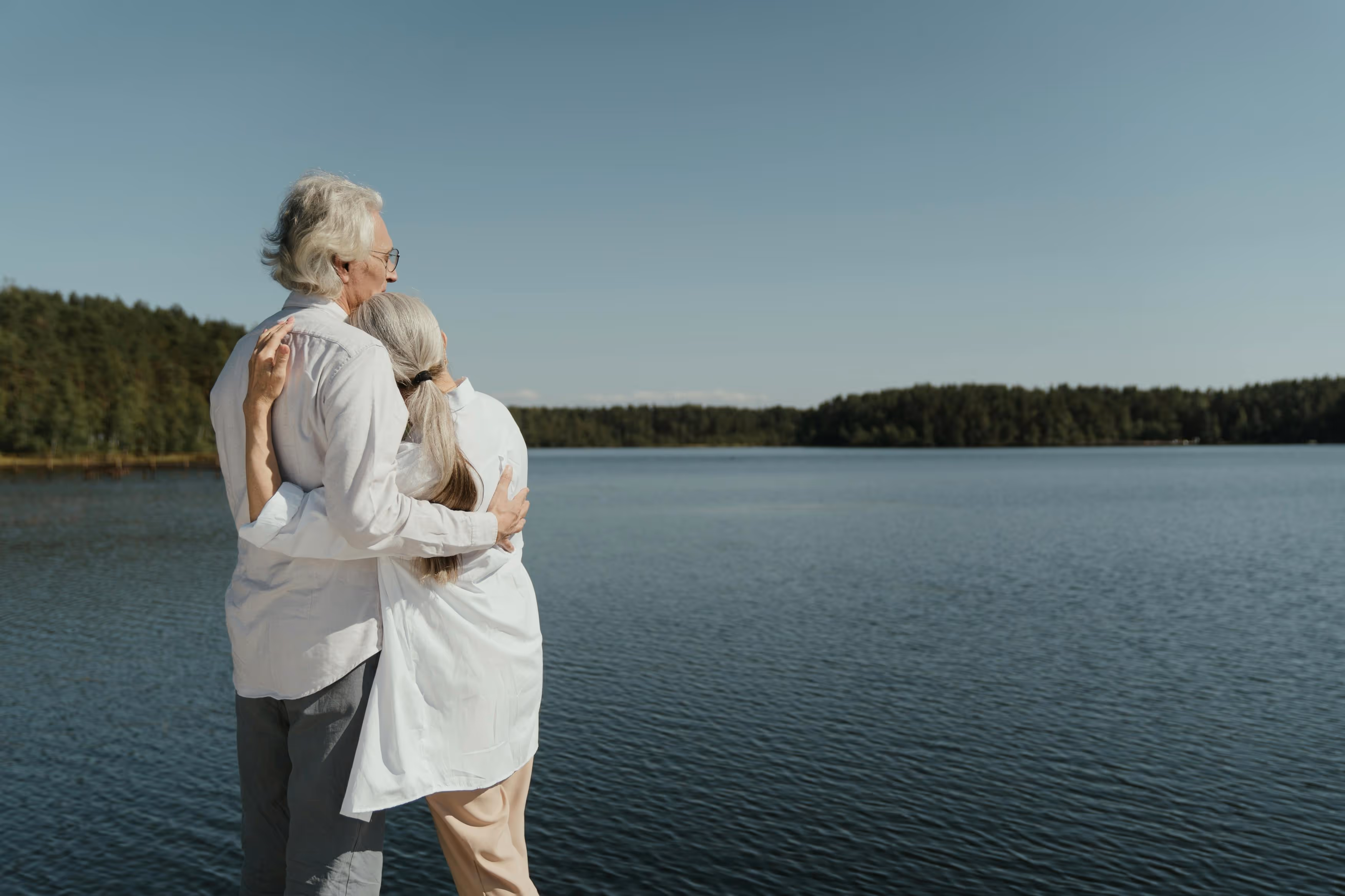 Elderly couple embracing while looking out over a calm lake with forested shores under a clear sky.