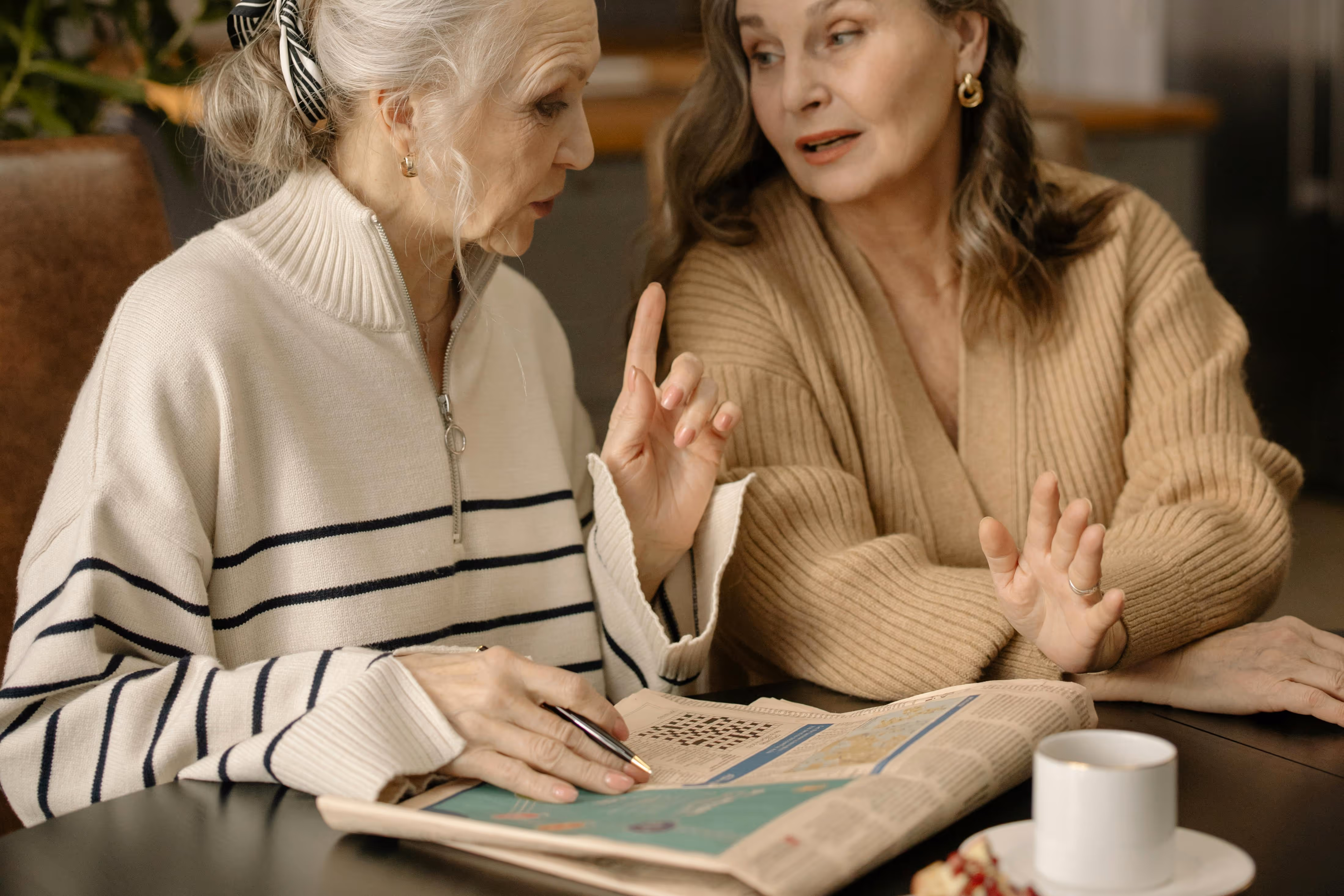 Two elderly women sitting at a table, one holding a pen and pointing while the other listens attentively, with a newspaper and a cup of coffee on the table.