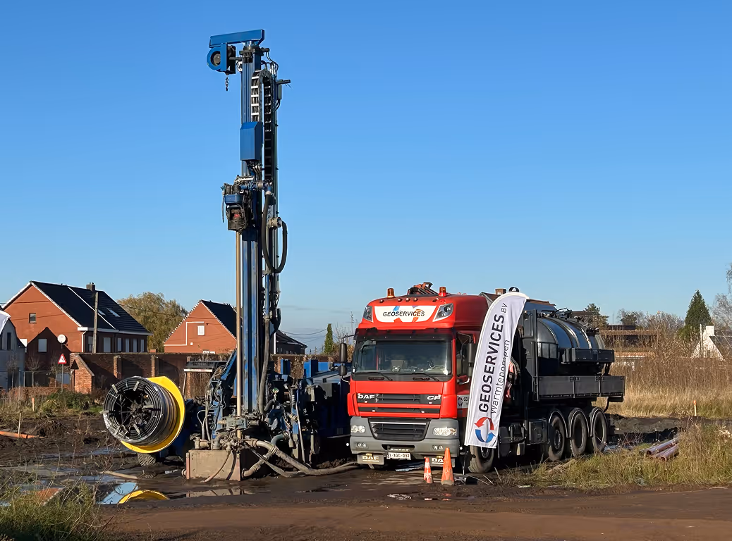 Een rode vrachtwagen van Geoservices naast een boortoren, met een helderblauwe lucht en huizen op de achtergrond.
