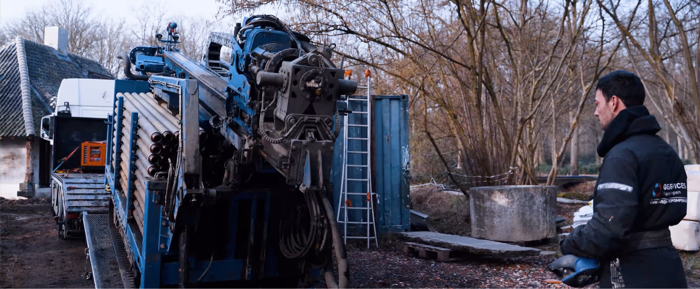Een werknemer in een jas staat naast een grote blauwe boormachine volgeladen met buizen in een bosrijke omgeving.
