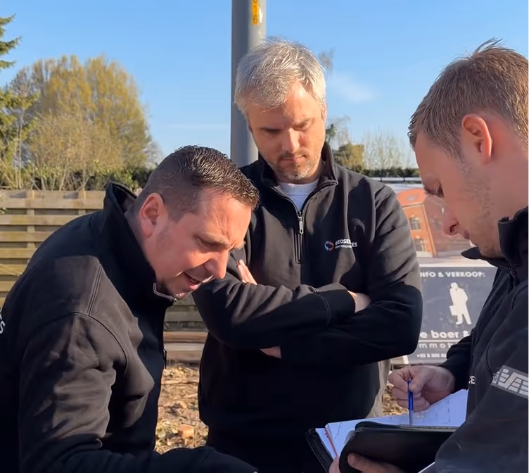 Drie mannen in zwarte jassen die op een zonnige dag buiten bij een houten schutting aantekeningen doornemen.