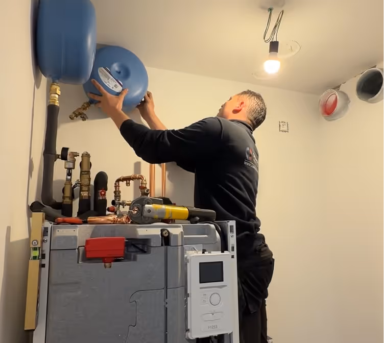 Een monteur die een blauw expansievat boven een verwarmingsinstallatie installeert of afstelt.