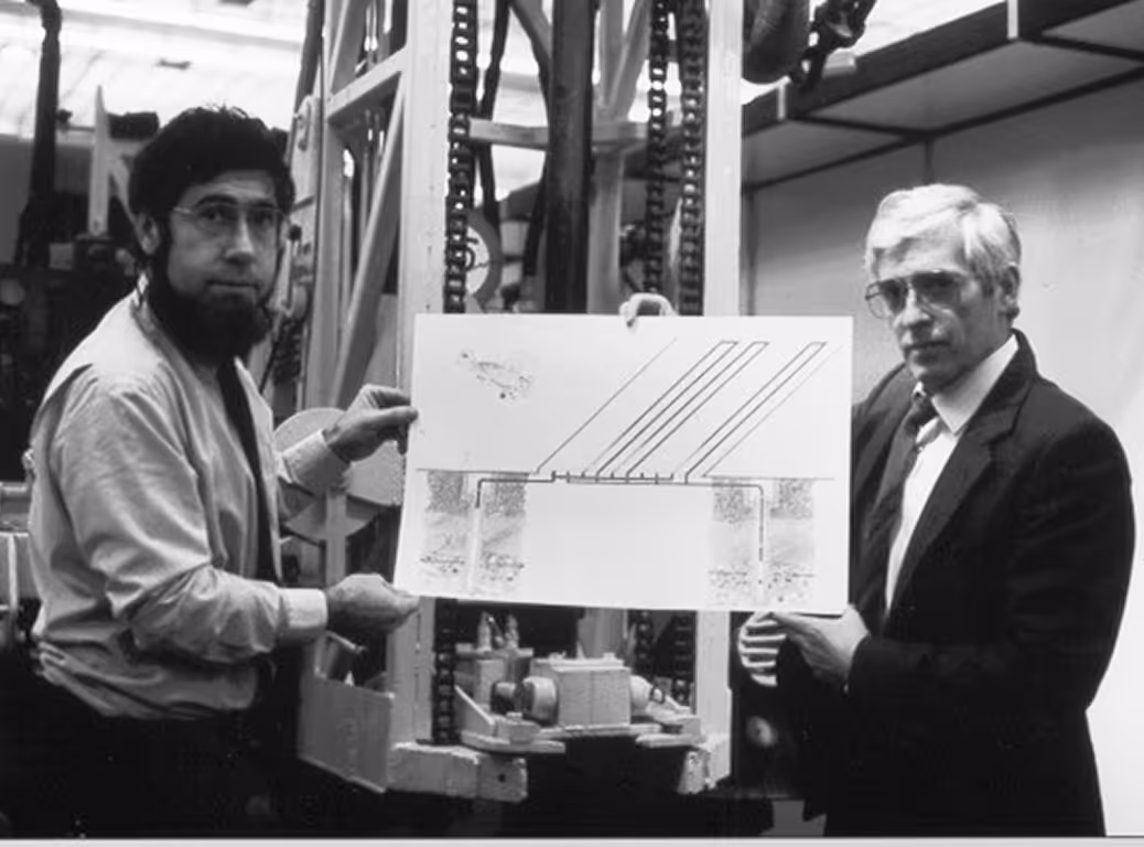 Twee mannen houden een technische tekening vast in een industriële omgeving, met machines op de achtergrond.