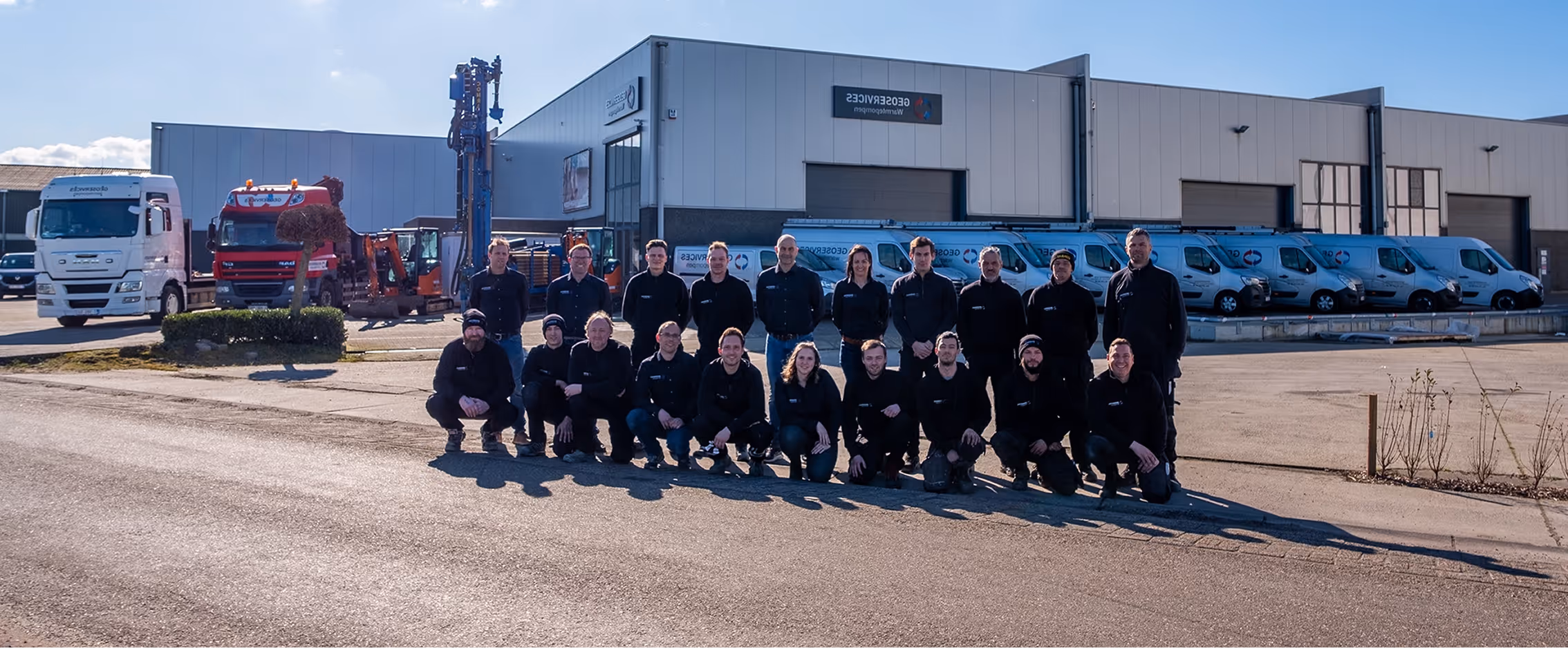 Een groep werknemers poseert voor een magazijn, met bestelwagens en vrachtwagens met bedrijfslogo’s die ernaast geparkeerd staan.