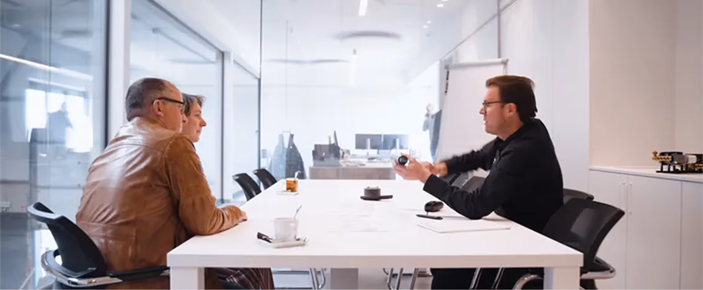 Een man met een bril legt een apparaat uit aan een man en een vrouw die aan weerszijden van een witte tafel zitten in een modern kantoor.