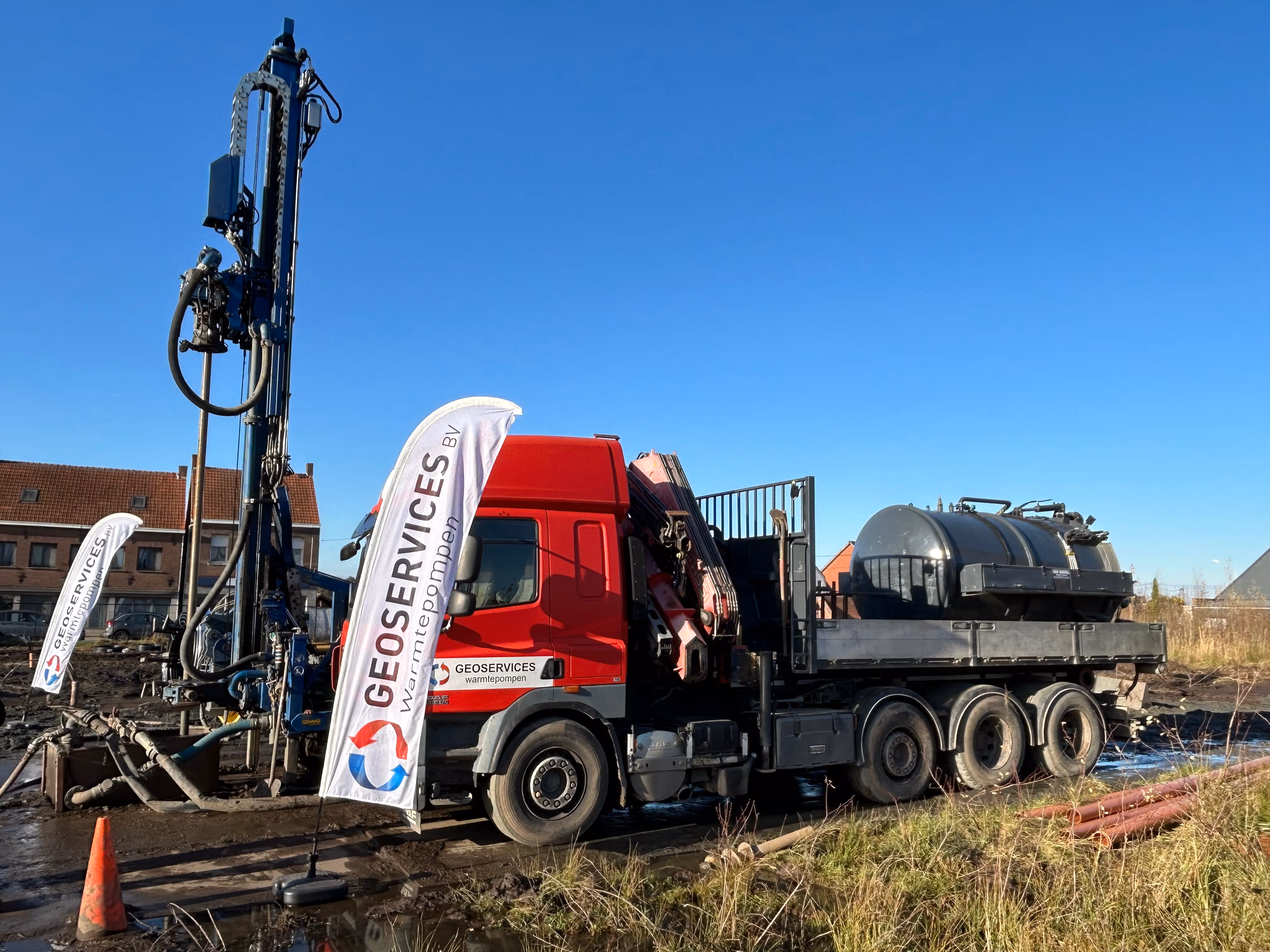 Een rode vrachtwagen van Geoservices met boorapparatuur op een modderige geothermische boorlocatie op een zonnige dag.