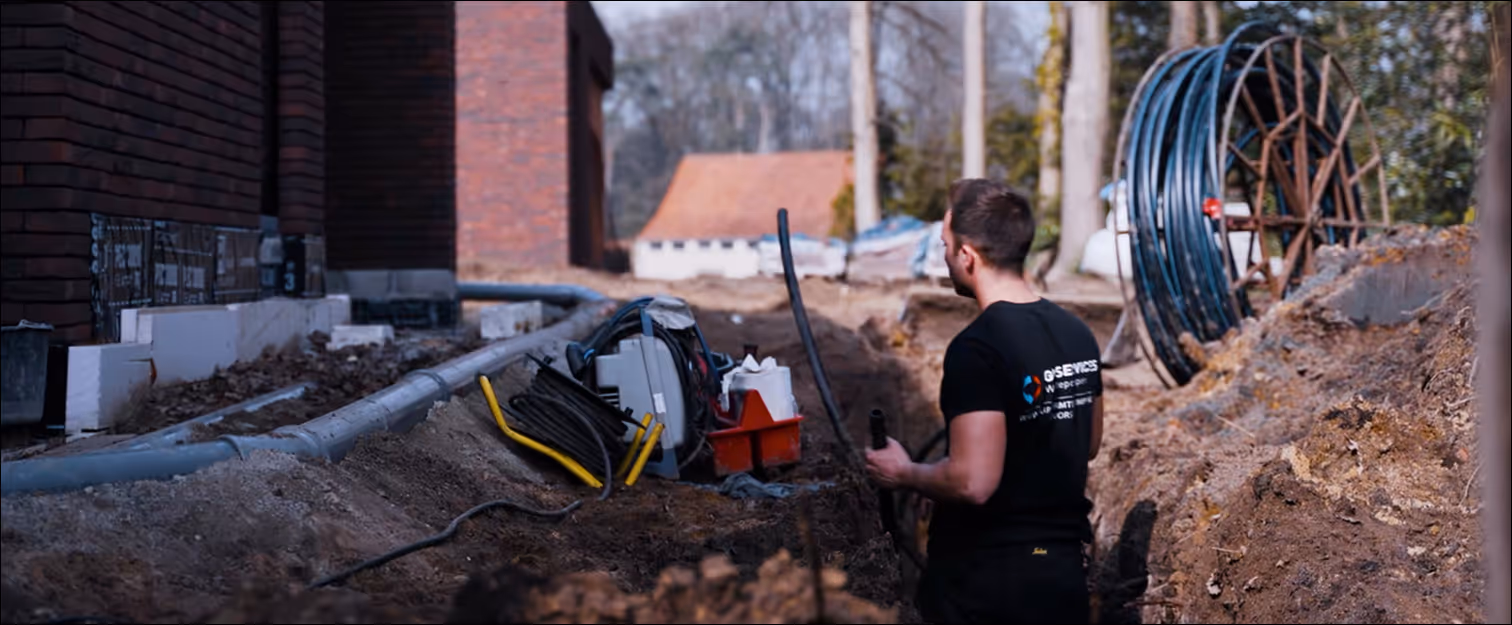 Een arbeider in een zwart shirt die kabels en apparatuur hanteert in een uitgegraven greppel naast een bakstenen gebouw.