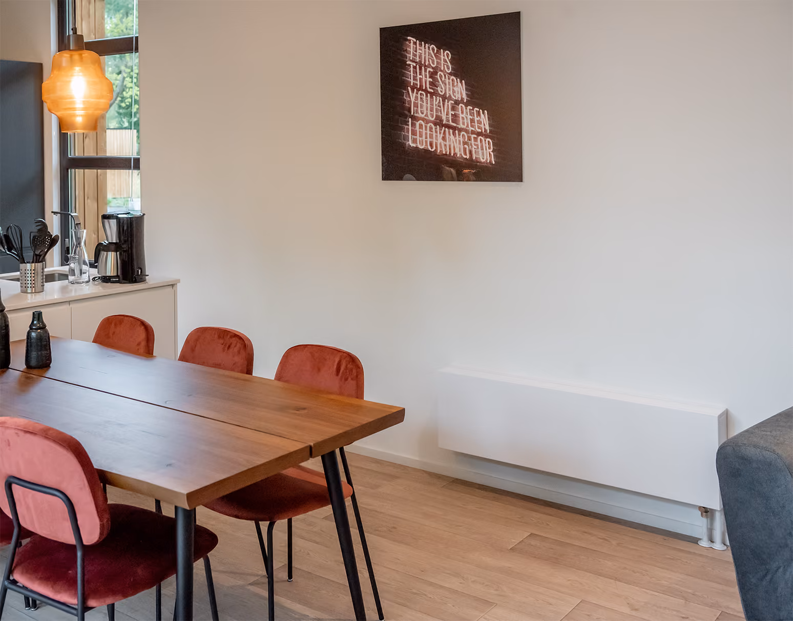 Eetkamer met houten tafel, vier rode stoelen, witte radiator en neonbord aan de muur.