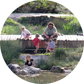 Un homme et quatre enfants jouent au bord d'un étang entouré de verdure et de rochers.