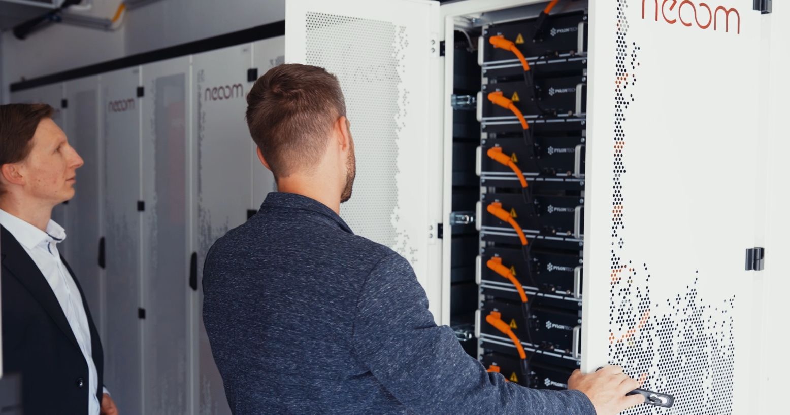 Two men inspecting an open server rack with orange cables connected to black equipment inside a white cabinet.