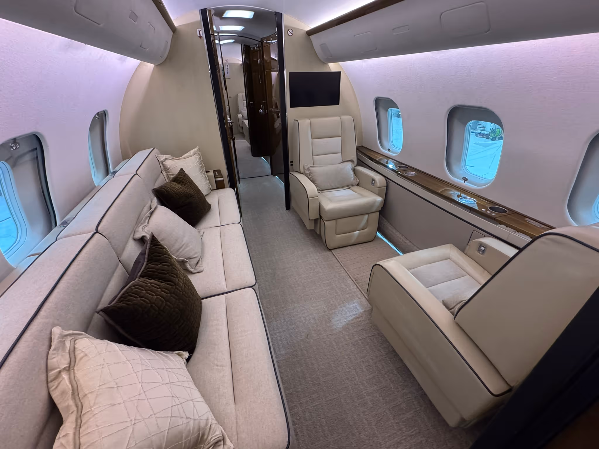 Luxurious private jet interior with beige leather seats, sofa with pillows, and windows along the side.