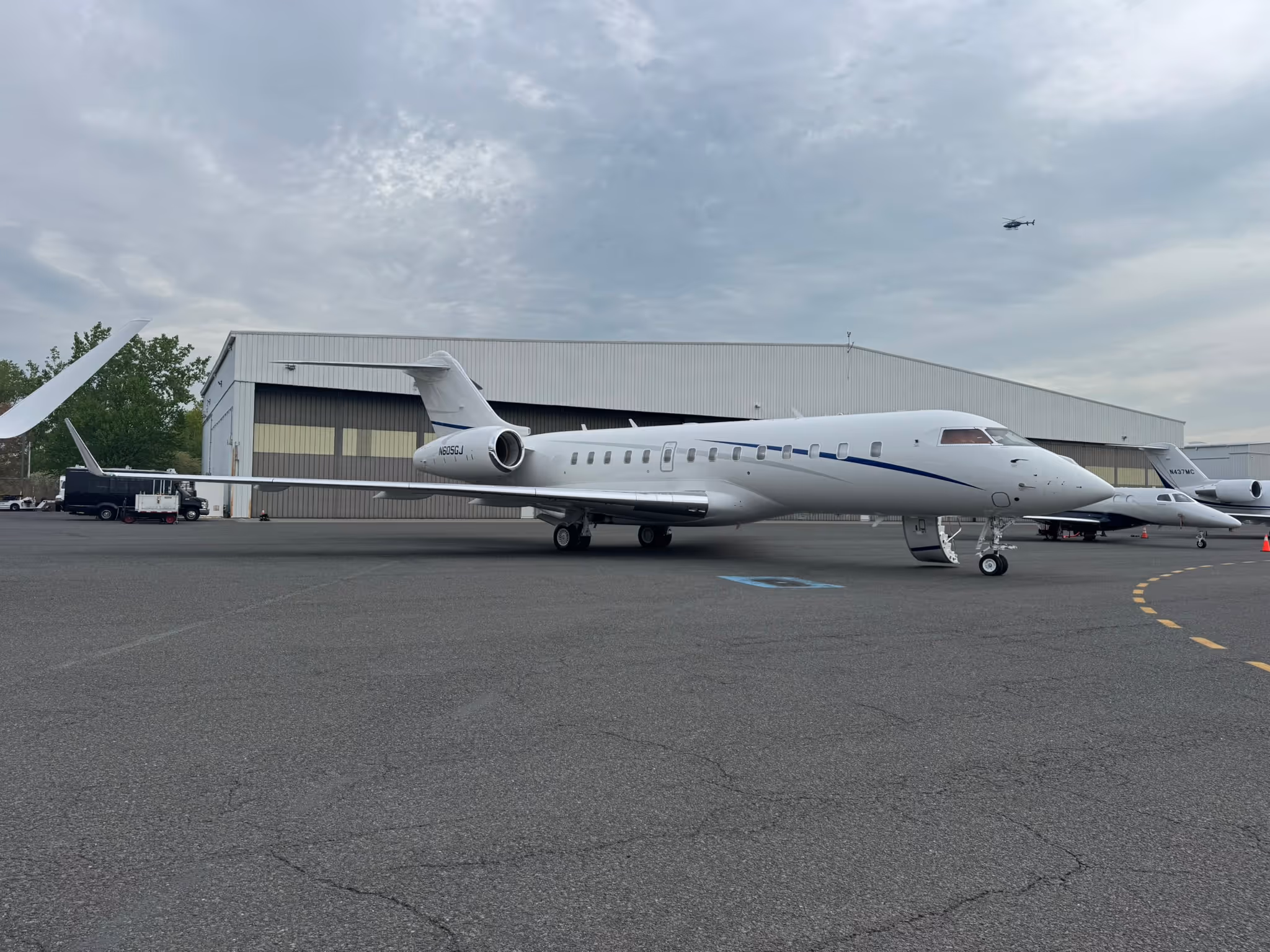White private jet with blue stripe parked on airport tarmac near hangars under cloudy sky.