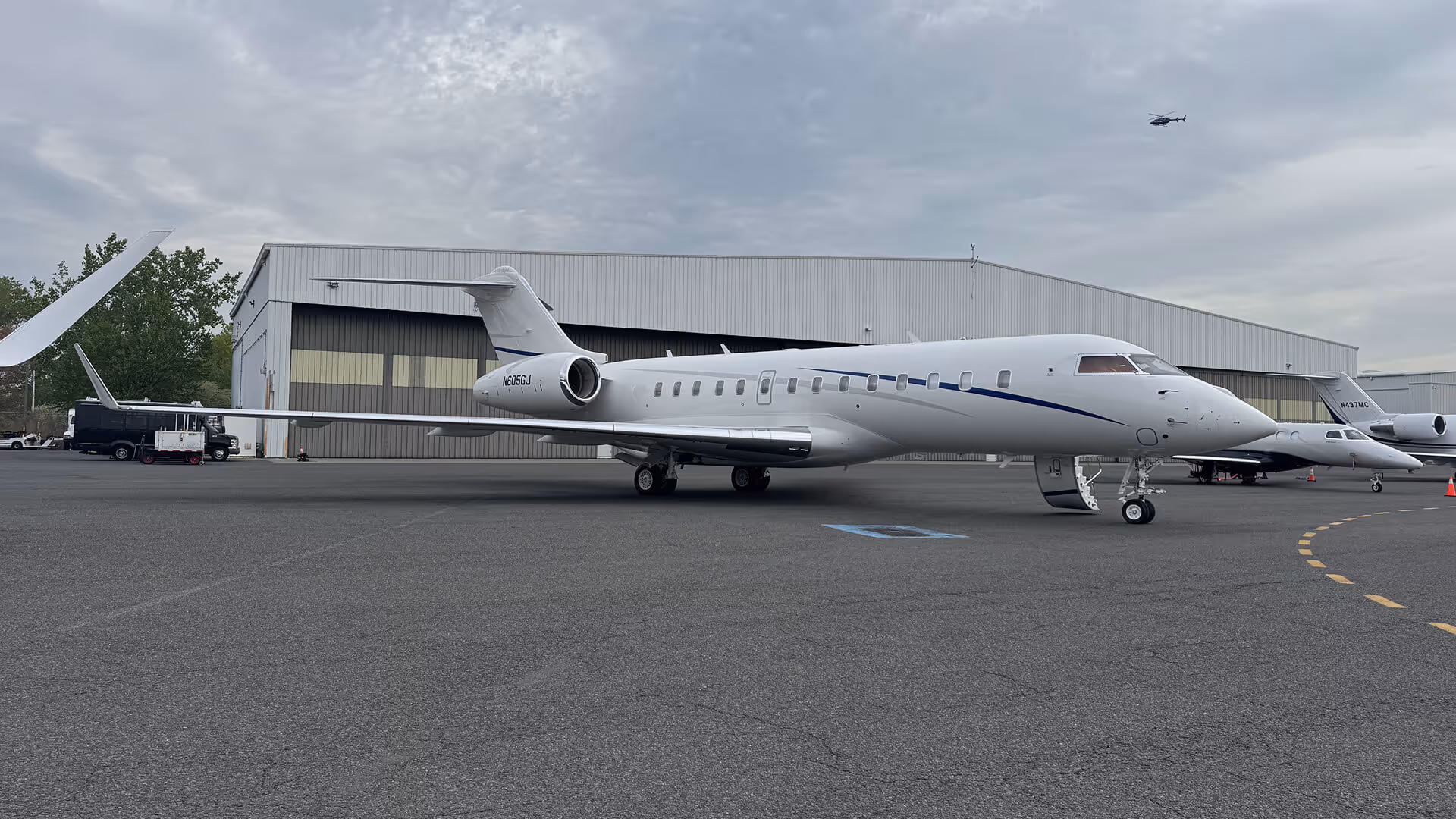 White private jet parked on tarmac near hangar with a helicopter flying in the cloudy sky above.