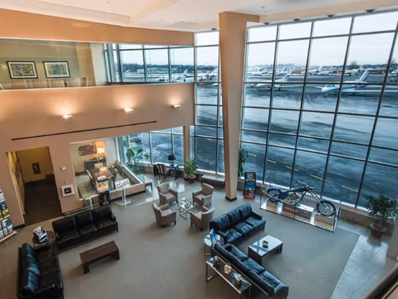 Modern airport lounge with seating, motorcycle display, and large windows overlooking parked planes.