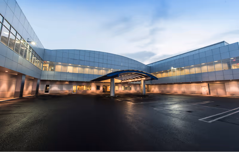 Modern building with large windows and a covered entrance in a mostly empty, wet parking lot at dusk.