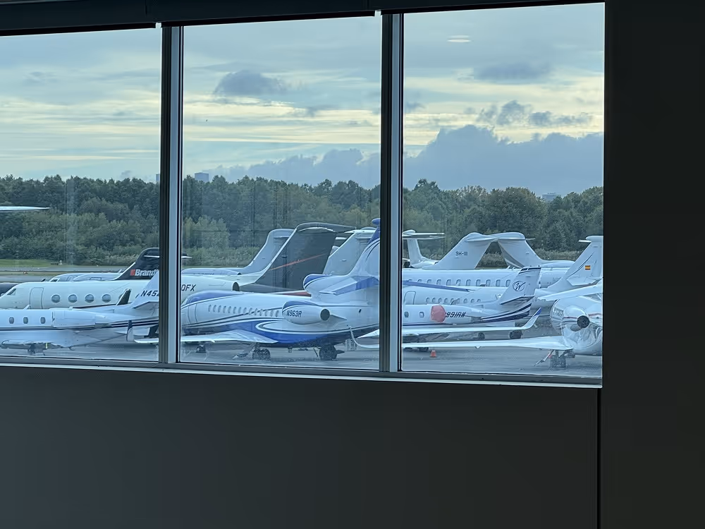 View through airport window of several parked small private jets against cloudy sky and trees.