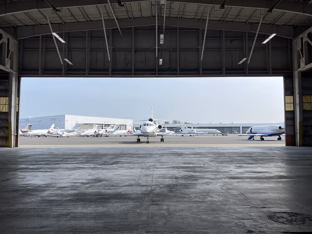 View of private jets parked outside on the airfield, seen from inside a large hangar.