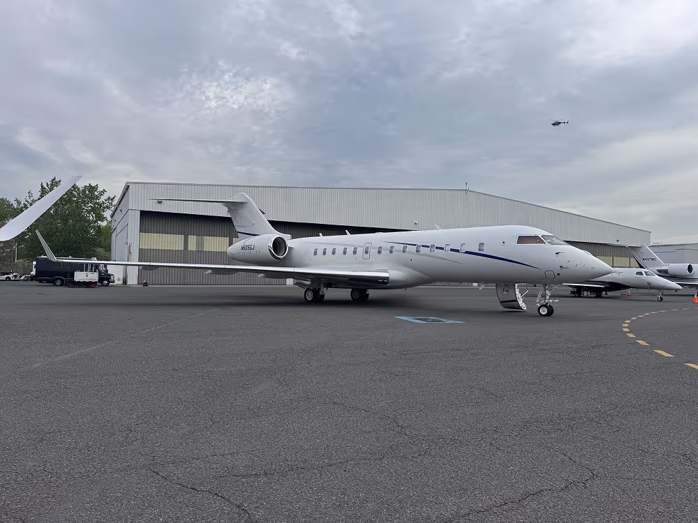 White private jet parked on an airstrip near a gray hangar under a cloudy sky with a helicopter flying above.