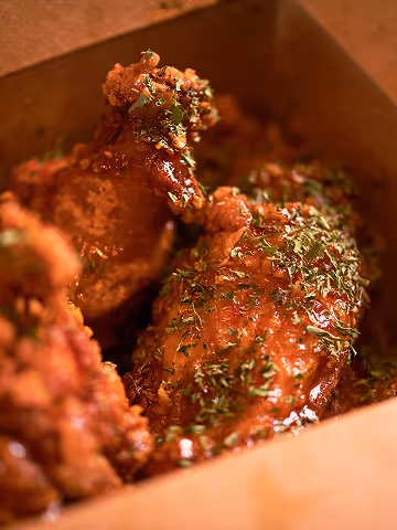 Close-up of glazed crispy chicken wings garnished with herbs in a brown takeout container.
