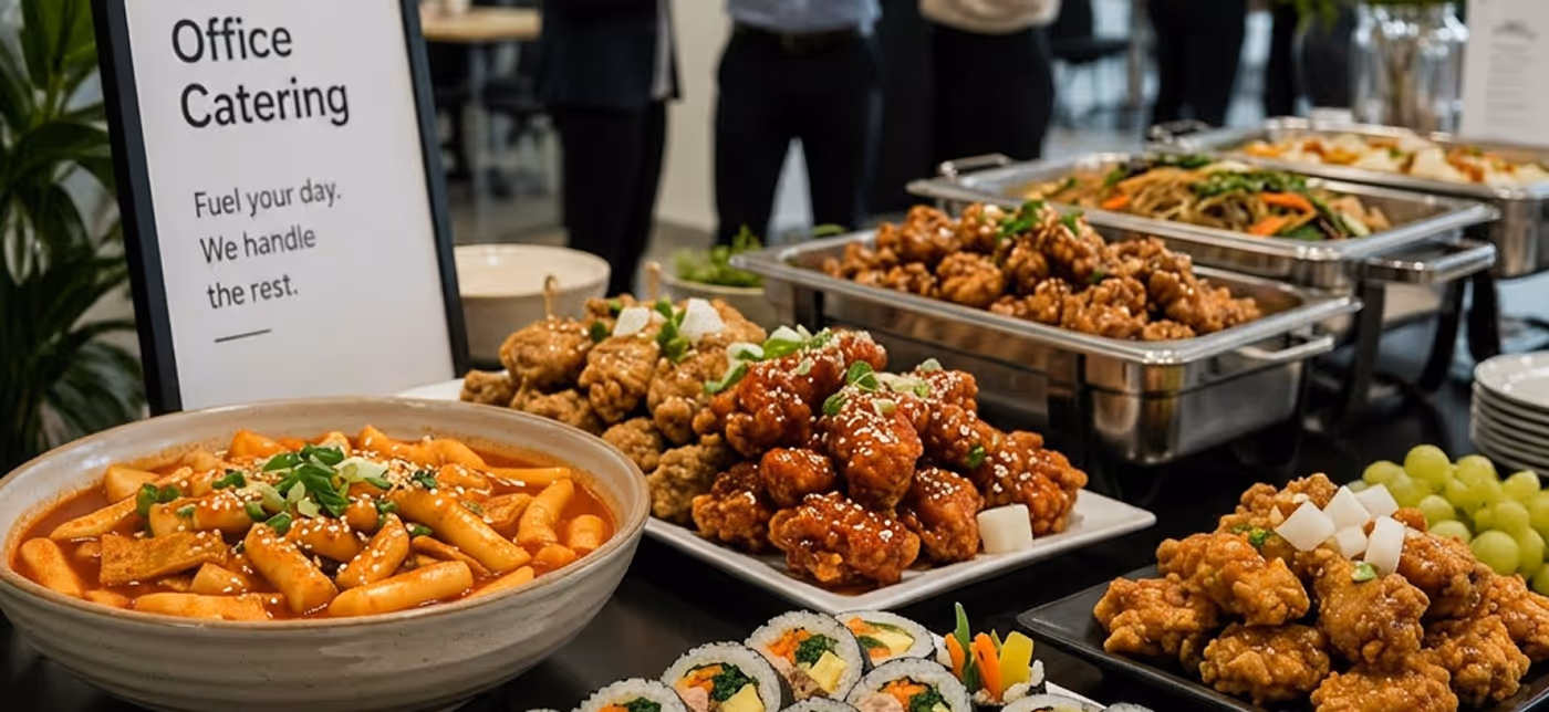 A variety of Korean food dishes including tteokbokki, fried chicken, sushi rolls, and grapes arranged on a buffet table with a sign for office catering.