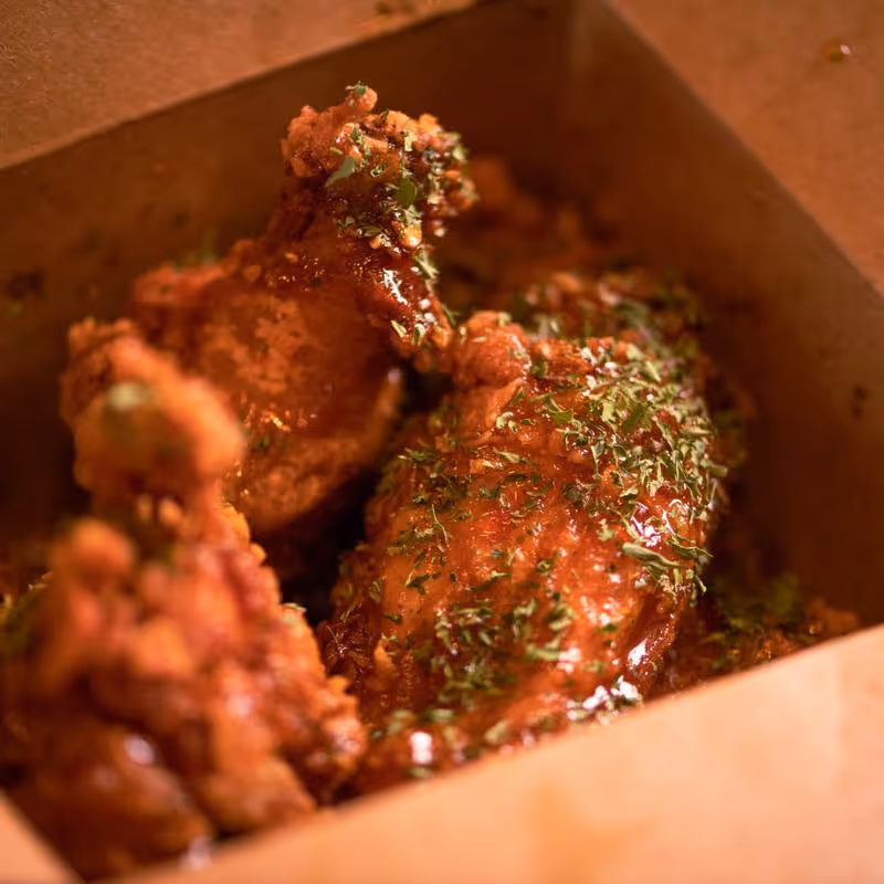 Close-up of crispy fried chicken wings coated in sauce and sprinkled with chopped herbs inside a cardboard box.