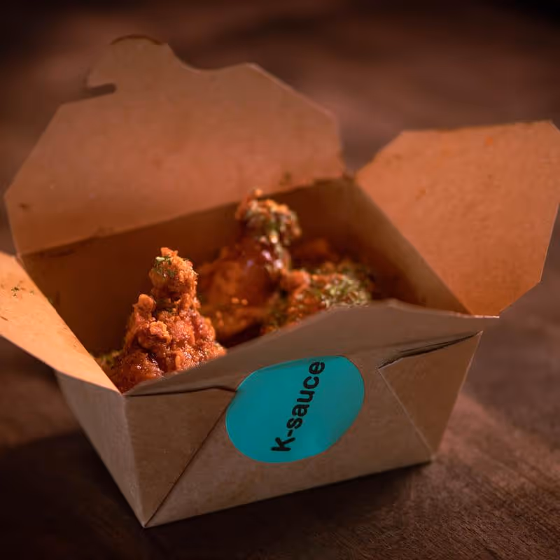 Takeout box with fried chicken pieces coated in sauce and sprinkled with herbs, labeled with a blue sticker saying 'k-sauce'.