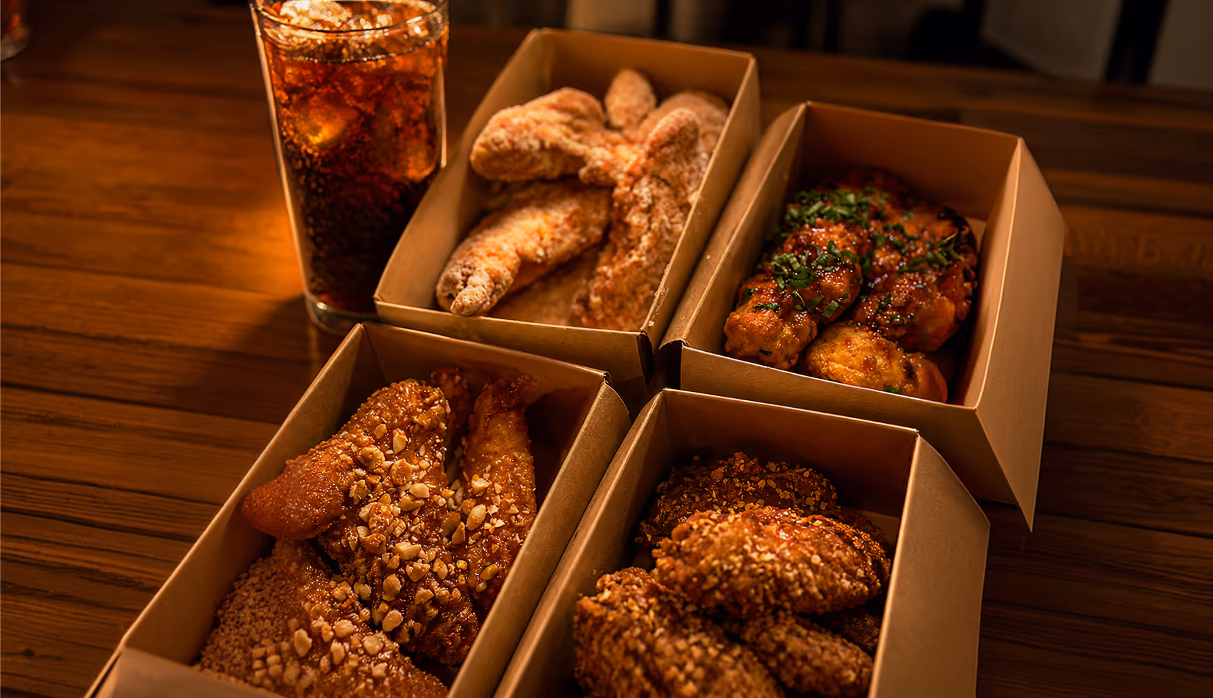 Four boxes of assorted fried chicken wings with different coatings and a glass of soda on a wooden table.