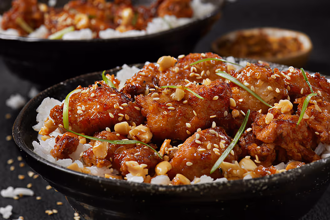 Bowl of glazed fried chicken pieces on white rice topped with sesame seeds, chopped nuts, and green onions.