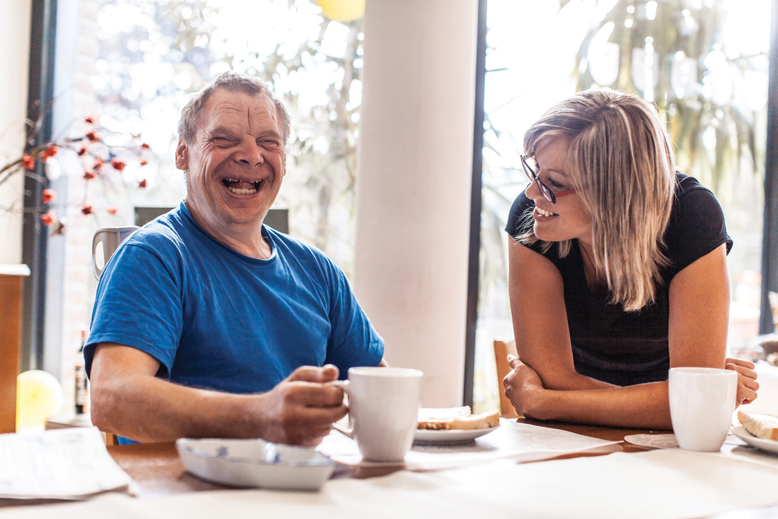 Man in a blue shirt laughing joyfully while holding a white mug, sitting at a table with a woman in glasses smiling at him.