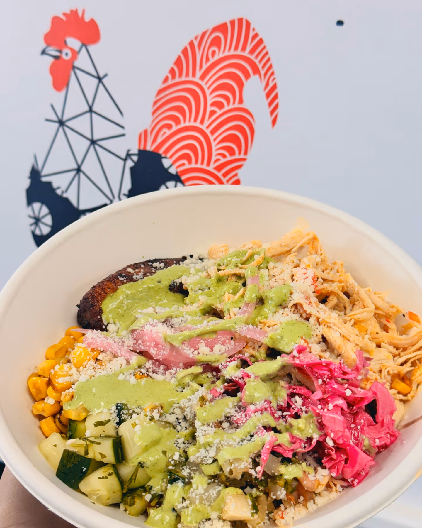 Bowl of shredded chicken, grilled corn, chopped cucumbers, pickled onions, pink cabbage, and grilled plantain topped with green sauce and crumbled cheese.
