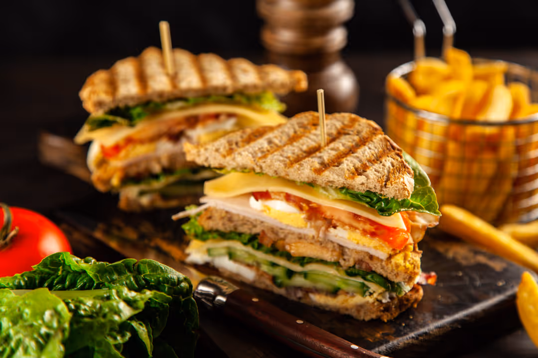 Grilled sandwich with lettuce, tomato, egg, cheese, and cucumber on a wooden board, served with a basket of French fries.
