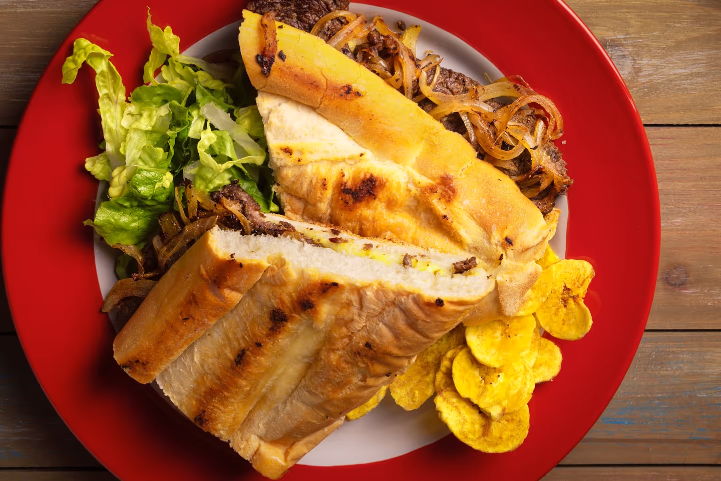 Grilled steak sandwich with caramelized onions, lettuce, and plantain chips on a red-rimmed plate.