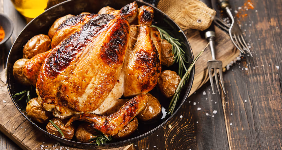 Roasted whole chicken with golden skin, surrounded by small roasted potatoes and rosemary in a black skillet on a wooden table.