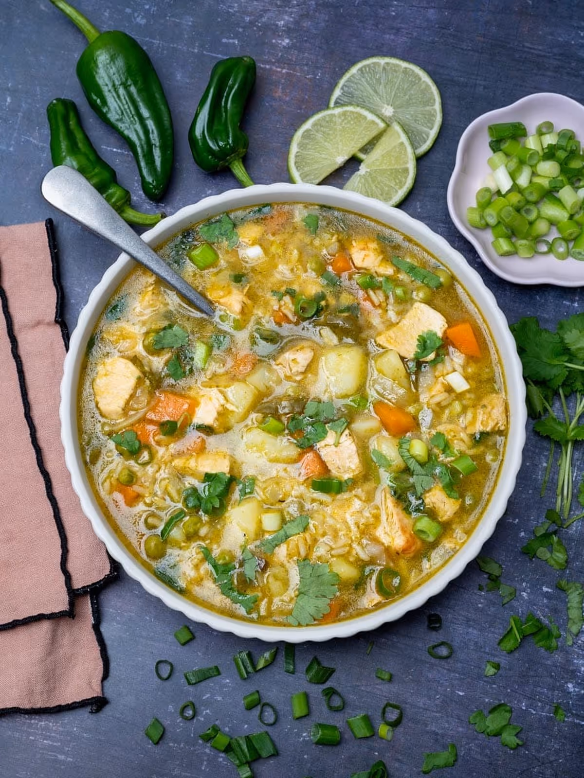 Bowl of chicken and vegetable soup garnished with green onions and cilantro, with sliced lime, whole green peppers, and chopped green onions on a dark surface.