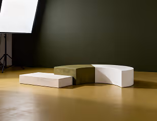 Three geometric pedestal blocks in beige, olive green, and white on a mustard floor against a dark green wall with a photo light reflector setup.