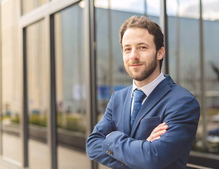 Confident man in a blue suit with arms crossed standing outside a modern glass building.