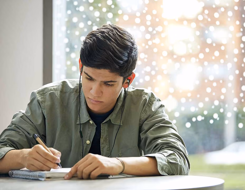 Young man wearing earphones writing in a notebook at a table with a window in the background.