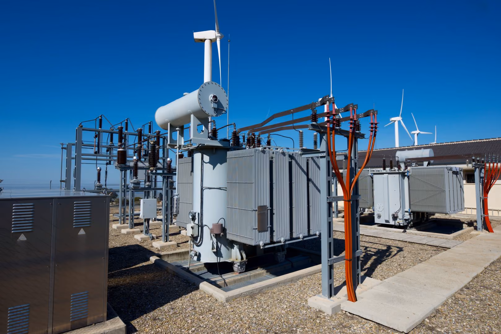 Electric substation with oil-filled transformers and wind turbines in the background under a clear blue sky.