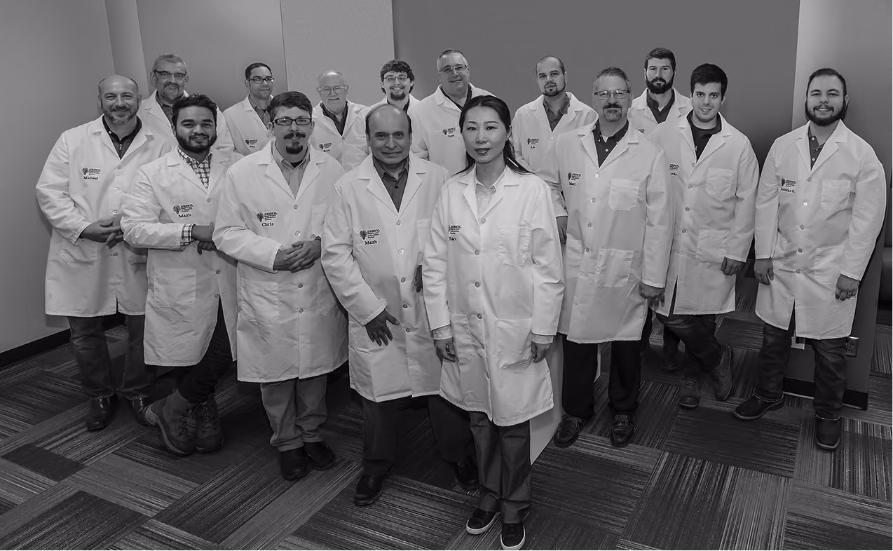 Group portrait of fifteen lab researchers wearing white coats standing in a room with carpeted floor.