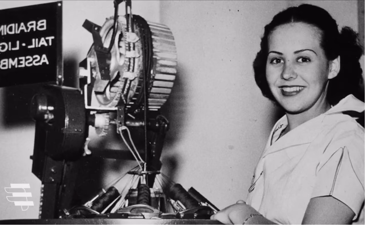 Smiling woman operating an early mechanical braiding machine in a black-and-white photo.